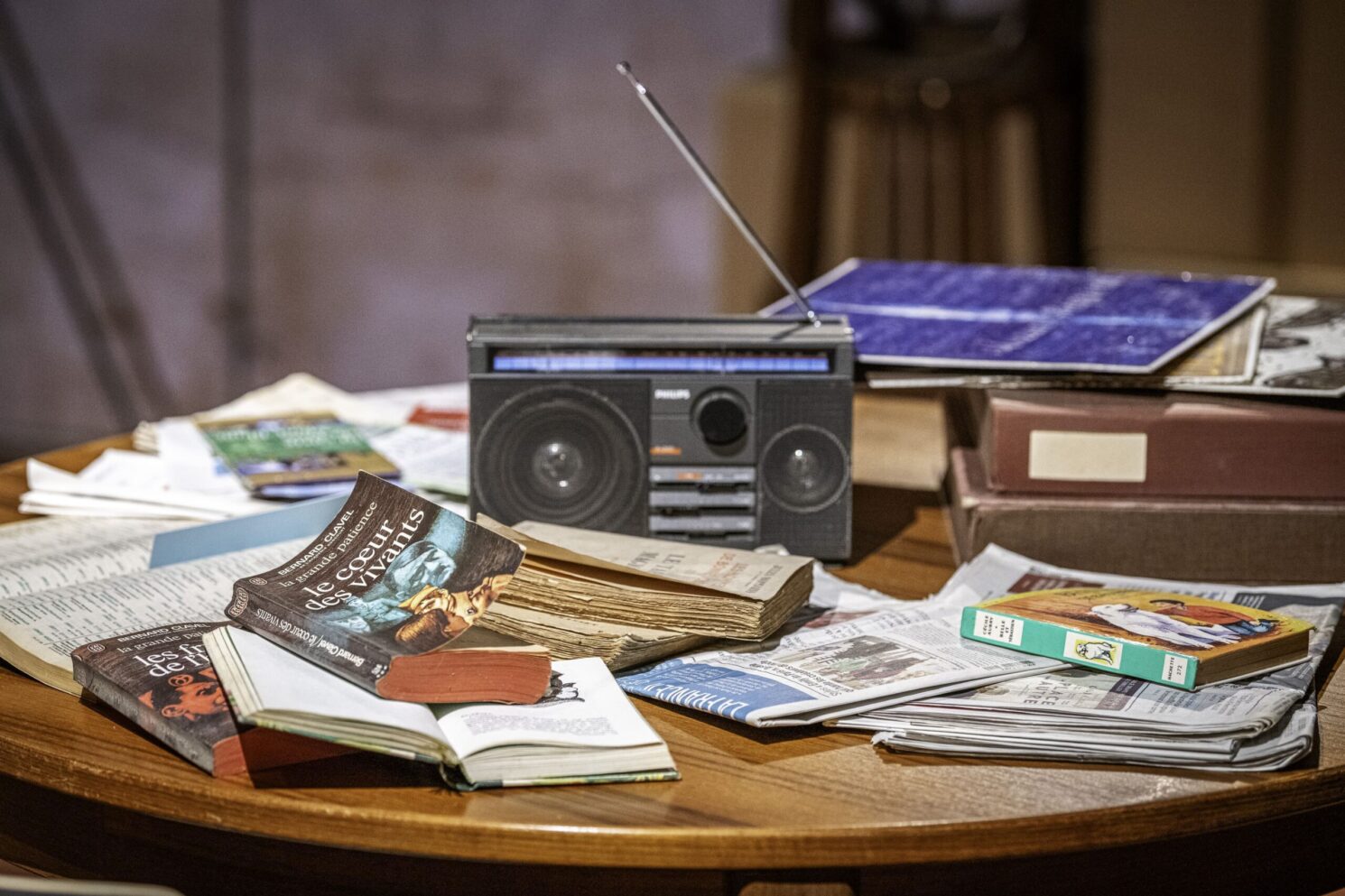 A curated still-life of everyday objects—including books, newspapers, a radio, and personal items—arranged on a table to recreate a familiar domestic environment, supporting the immersive storytelling and awareness-driven narrative of the Action Innocence traveling educational pavilion.