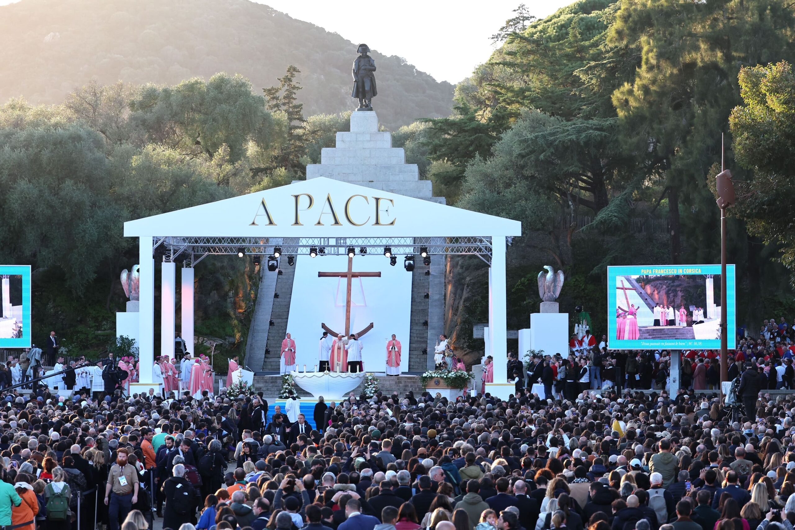A wide, panoramic view of a large-scale outdoor religious ceremony held during Pope Francis’ visit to Corsica. The monumental stage is set against a hillside and stone monument, crowned with the words “A PACE” in large gold lettering. At the center of the stage stands a tall wooden cross behind an altar shaped like a boat, surrounded by senior clergy members in pink and white liturgical vestments. Large LED screens on either side broadcast the ceremony to the crowd. In the foreground, a vast audience gathers closely together, emphasizing the scale and significance of the event. Professional stage lighting, floral arrangements, and the dramatic natural backdrop combine to create a solemn, historic, and highly symbolic atmosphere characteristic of a major papal public appearance.