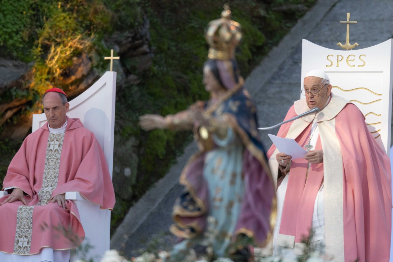 A wide outdoor shot of a religious address being delivered. The speaker stands behind a lectern, reading from a document, while another clergyman sits nearby. A religious statue is visible in the foreground, slightly out of focus, adding depth and context to the scene.