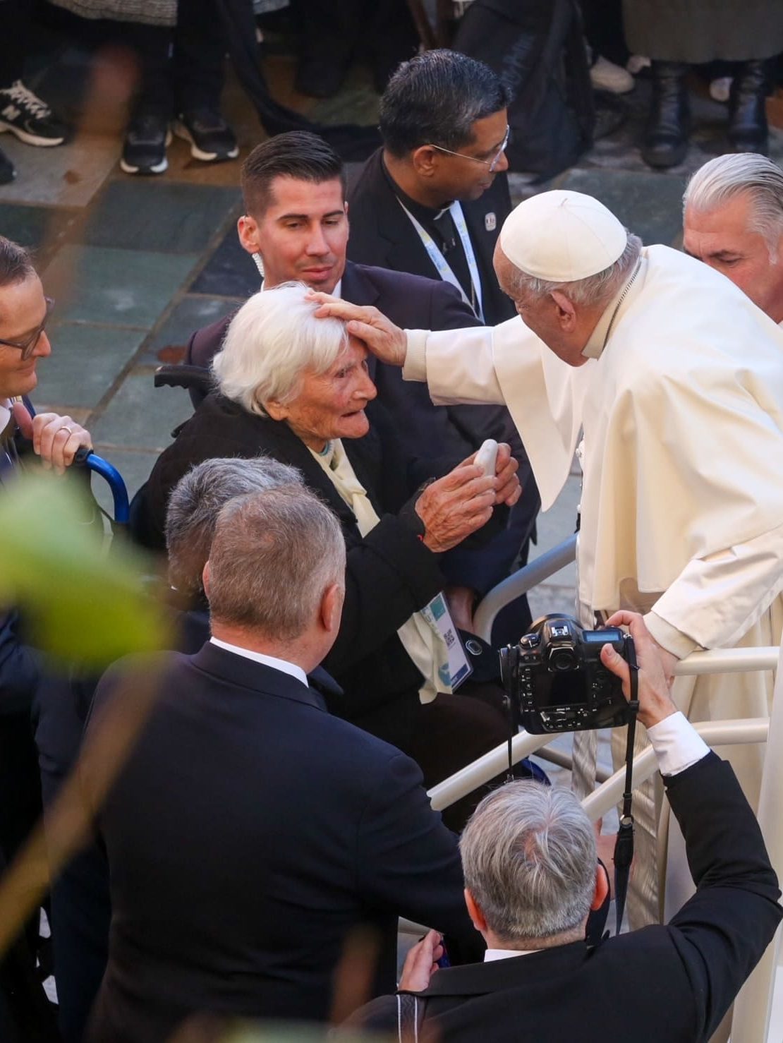 An overhead scene capturing a religious leader greeting and blessing an elderly woman among a small crowd. Several people surround them, including security and photographers, emphasizing the public and symbolic nature of the moment.