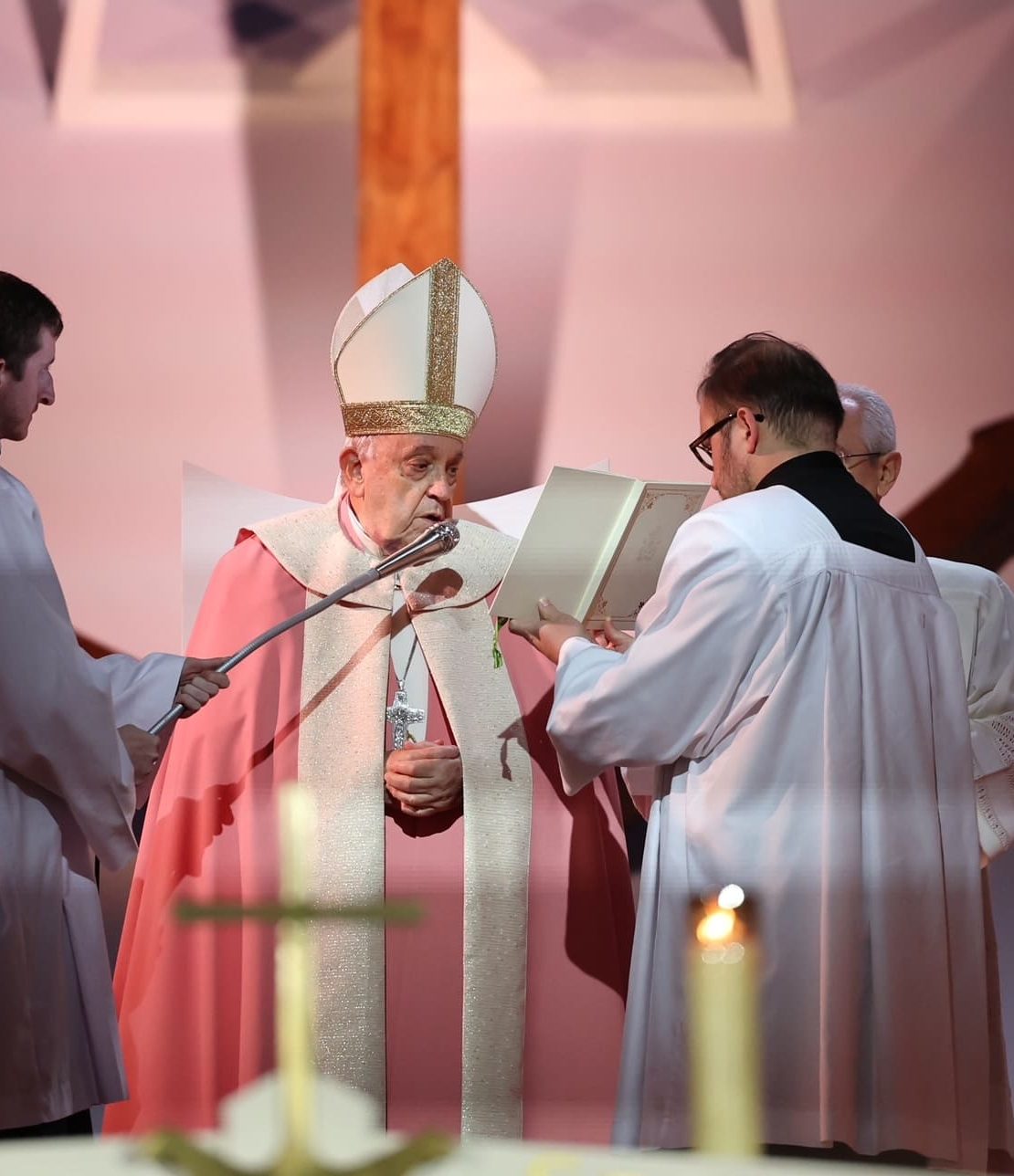 A wide view of a solemn religious ceremony during Pope Francis’ visit to Corsica. Pope Francis stands at the altar, seated beneath a large wooden cross, while another senior clergyman addresses him from a lectern. Soft stage lighting, lit candles in the foreground, and a minimalist backdrop reinforce the contemplative and ceremonial atmosphere of the event.