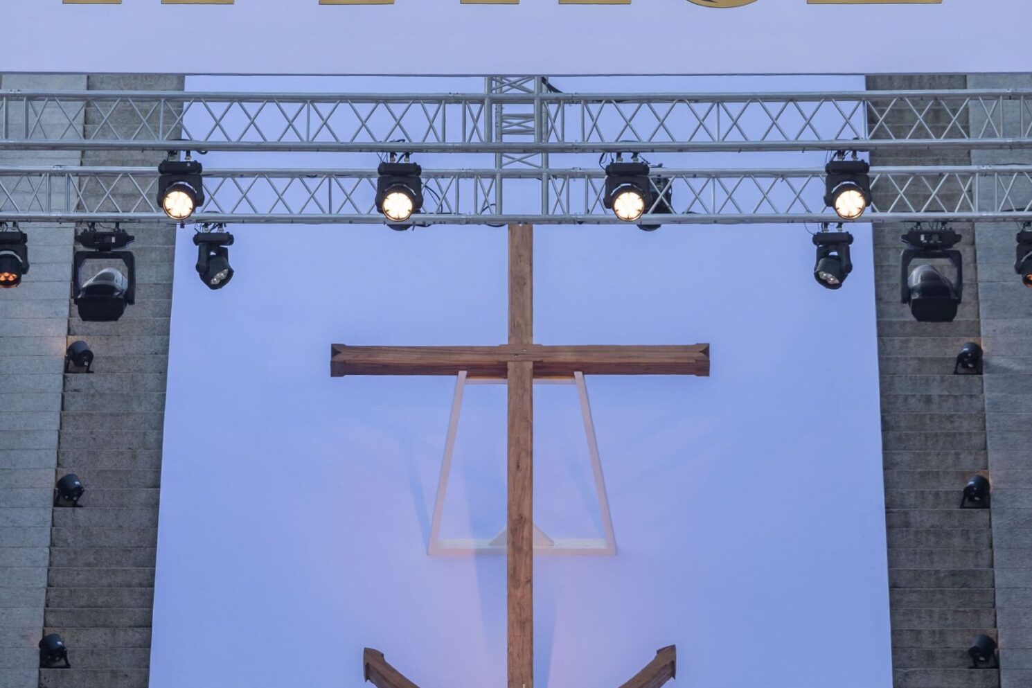 A large-scale outdoor religious ceremony set against a monumental stone backdrop, taking place during Pope Francis’ visit to Corsica. A prominent stage structure features the word “A PACE” in large gold lettering above a central wooden cross. Below, senior clergy members in ceremonial vestments are seated and standing around an altar shaped like a boat, addressing a large gathered crowd in the foreground. Professional stage lighting and floral arrangements frame the scene, conveying a solemn, formal, and highly symbolic atmosphere characteristic of a major public religious event.