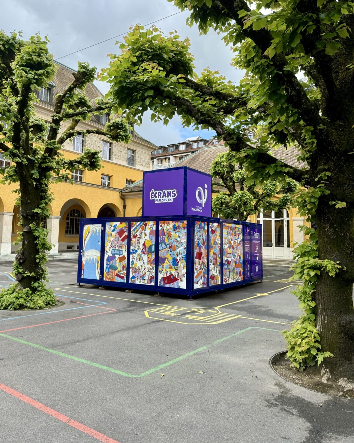 The Action Innocence traveling educational pavilion installed in an outdoor school courtyard, featuring colorful illustrated panels and branded signage, integrated into the environment beneath surrounding trees.