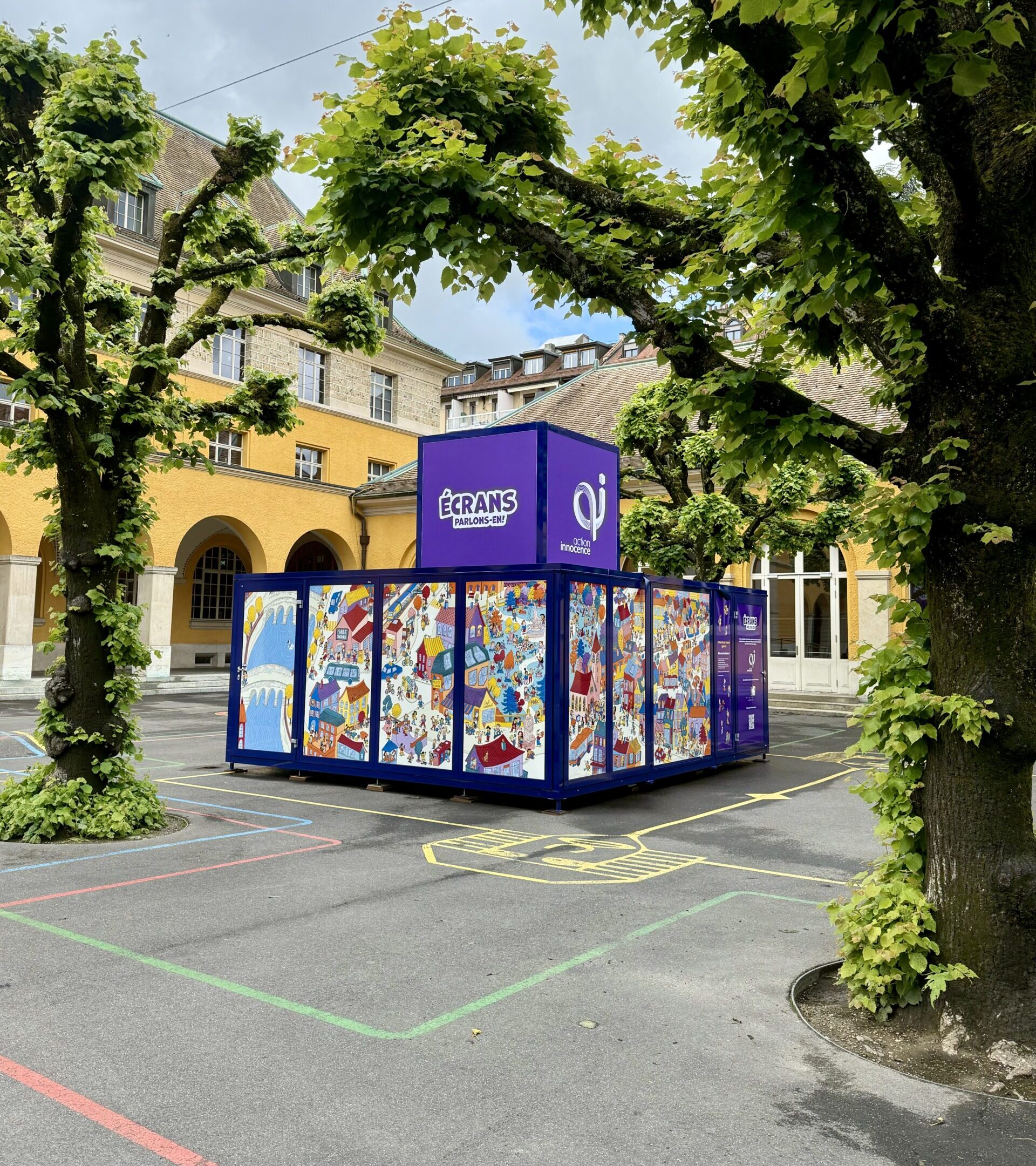The Action Innocence traveling educational pavilion installed in an outdoor school courtyard, featuring colorful illustrated panels and branded signage, integrated into the environment beneath surrounding trees.