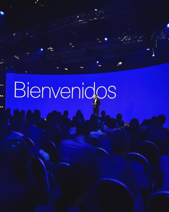 Speaker welcoming the audience on stage in front of a large blue screen reading “Bienvenidos” during the Conference in Buenos Aires – Immersive 360°.