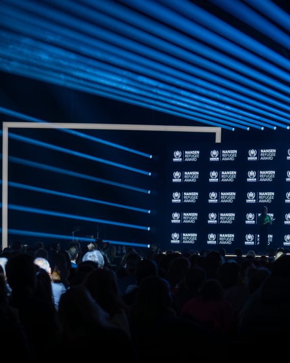 Wide view of the stage and audience during the Nansen Refugee Award – Hybrid Ceremony, with blue light beams, a branded UNHCR backdrop, and speakers addressing the crowd.
