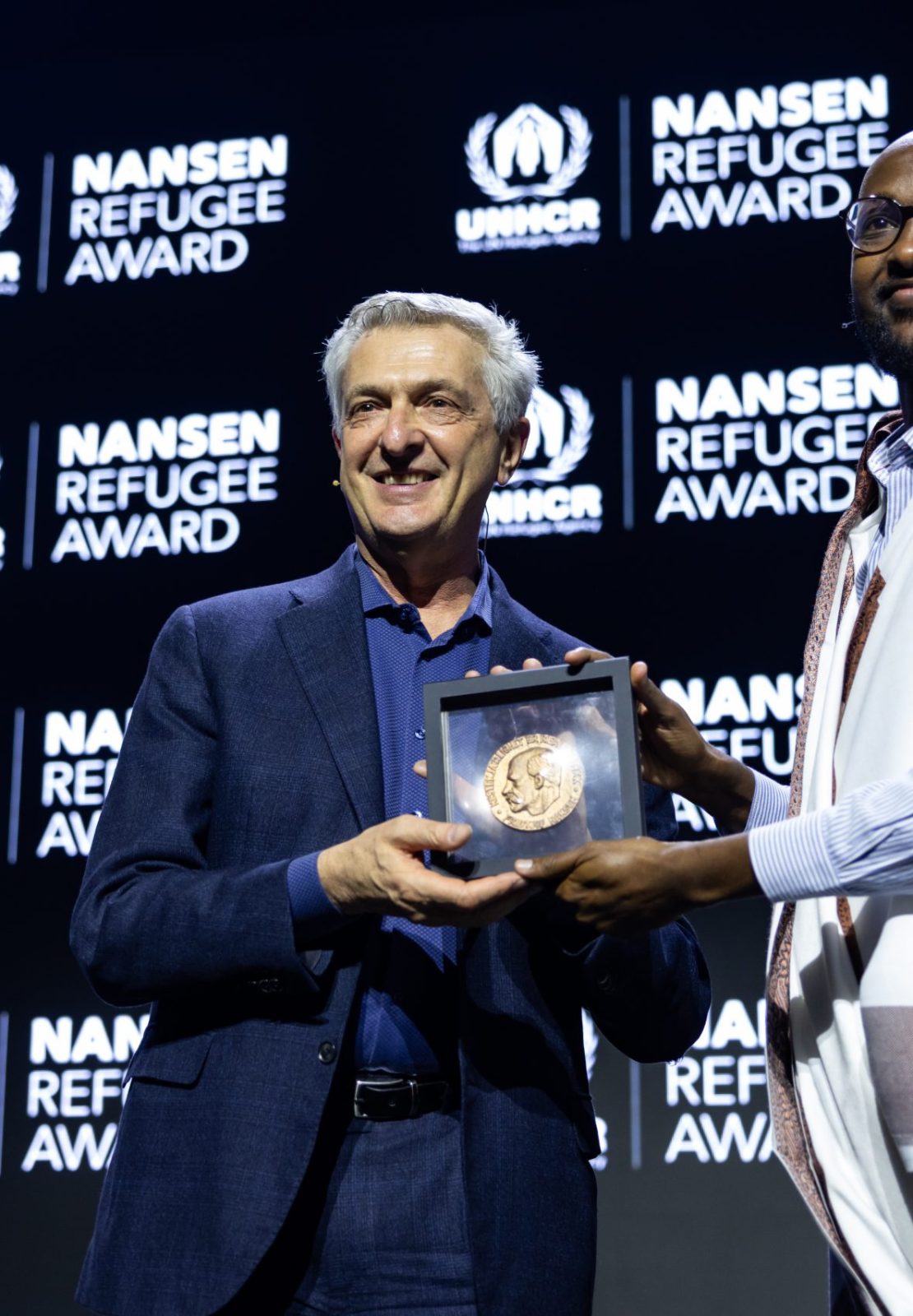 Two men holding an award together on stage during the Nansen Refugee Award – Hybrid Ceremony, standing in front of a backdrop displaying the UNHCR Nansen Refugee Award logo.
