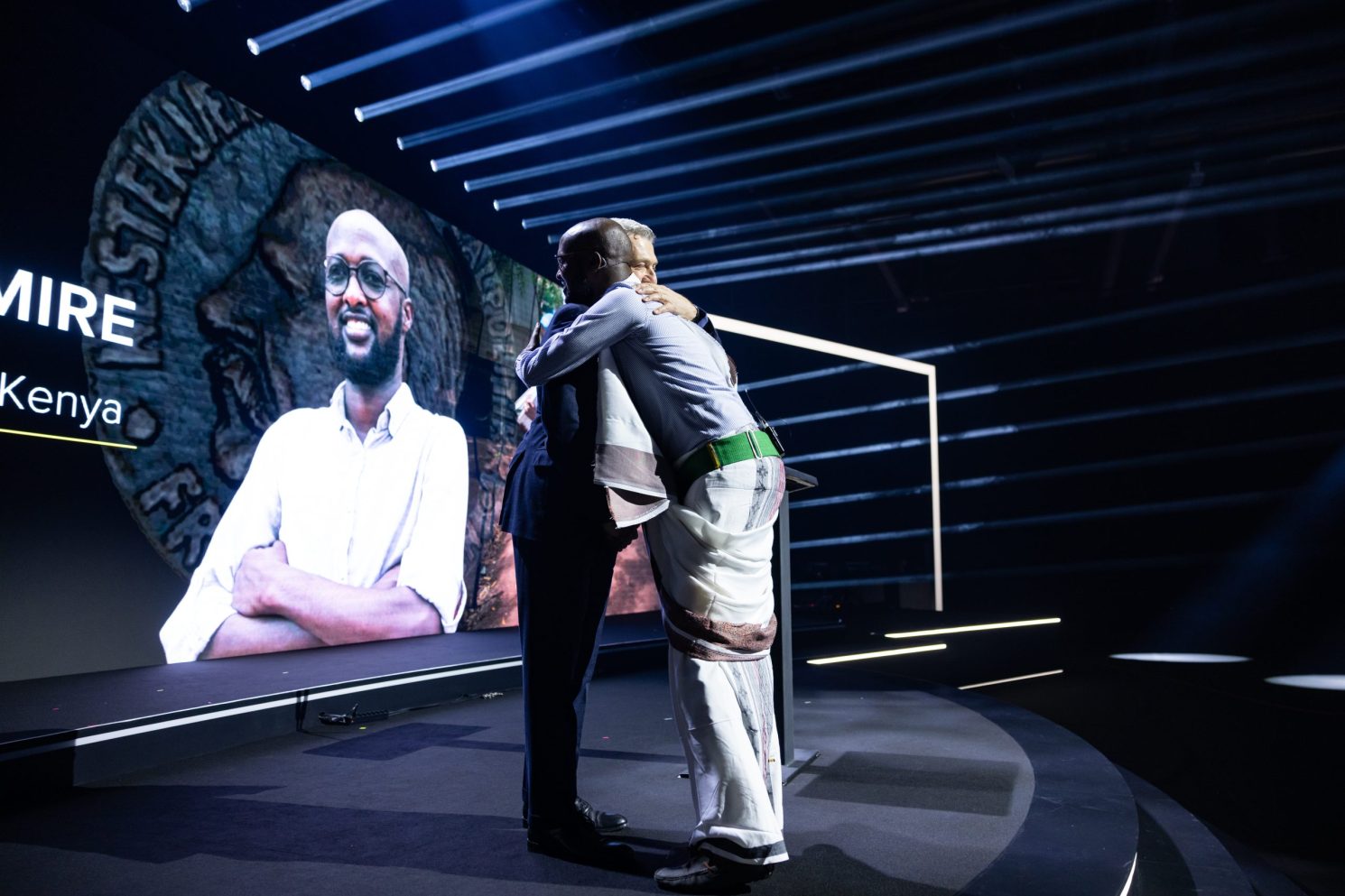 Two men embracing on a stage during the Switzerland 2023 UNHCR Nansen Refugee Award ceremony, with dramatic lighting and a large screen behind them displaying a portrait and text.