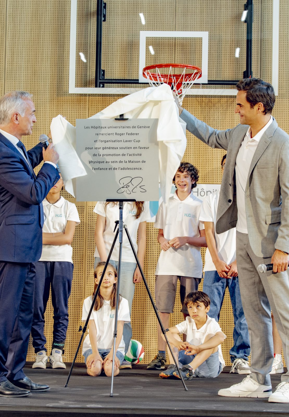 Two men unveiling a commemorative plaque during an official ceremony at the MEA – Opening of MEA at Geneva, on a stage surrounded by children and staff in a gymnasium setting.