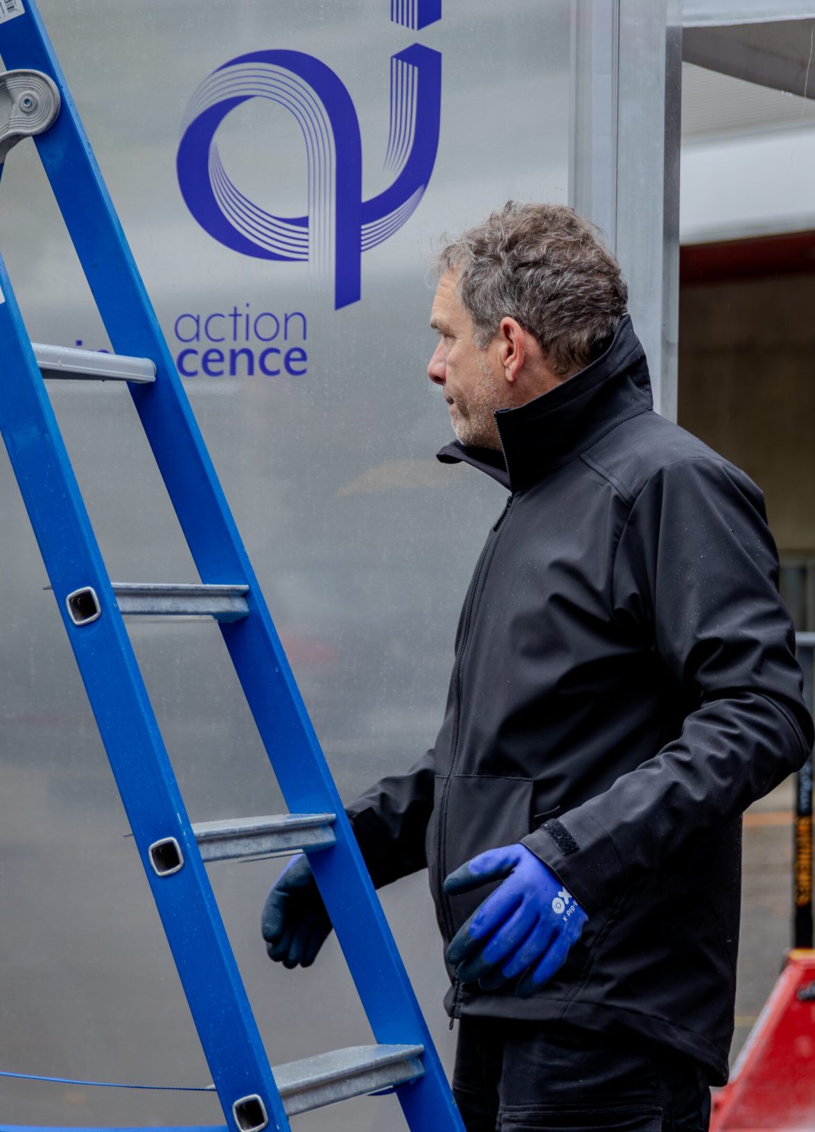 A technician prepares the installation of the Action Innocence traveling educational pavilion, standing beside a ladder in front of a translucent panel featuring the Action Innocence logo during the setup phase.